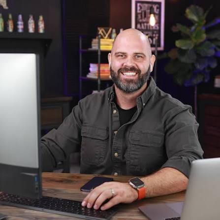 A man with a beard smiles at a desk with a computer and laptop, surrounded by shelves with books and bottles.