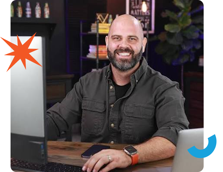A man with a beard smiles at a desk with a laptop, phone, and watch. A starburst and blue shape are overlaid.
