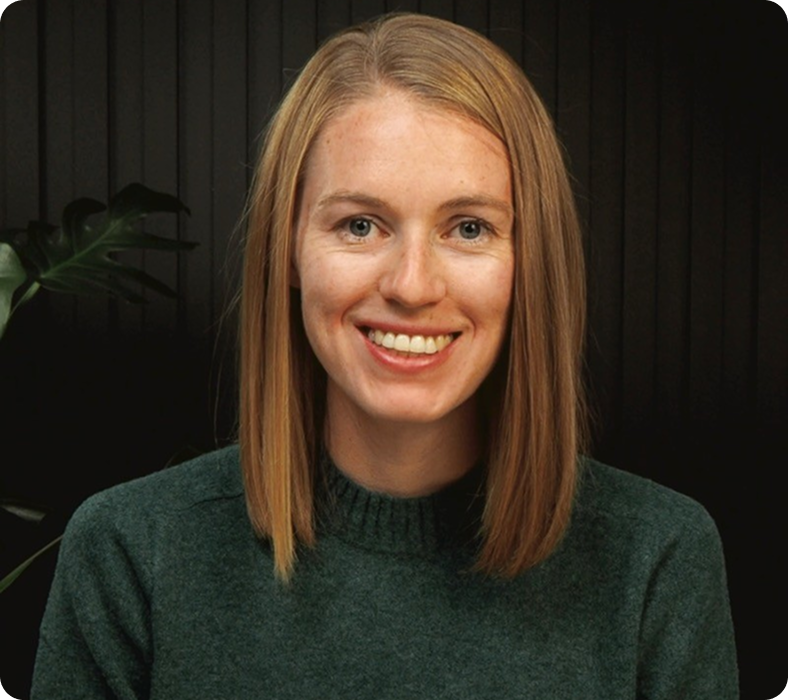 A smiling person with shoulder-length blonde hair wearing a dark green sweater stands against a dark background with a plant partially visible on the left side.