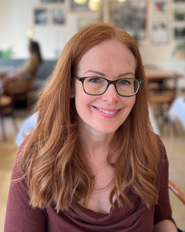 A woman with long red hair and glasses smiles in a softly lit room with blurred background elements.